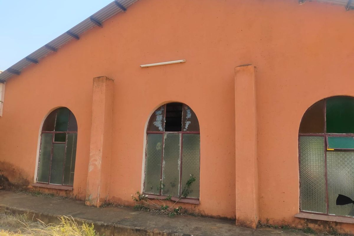 Exterior view of a commercial building featuring arched windows, a corrugated metal roof, and a concrete walkway, showing signs of significant wear and damage.