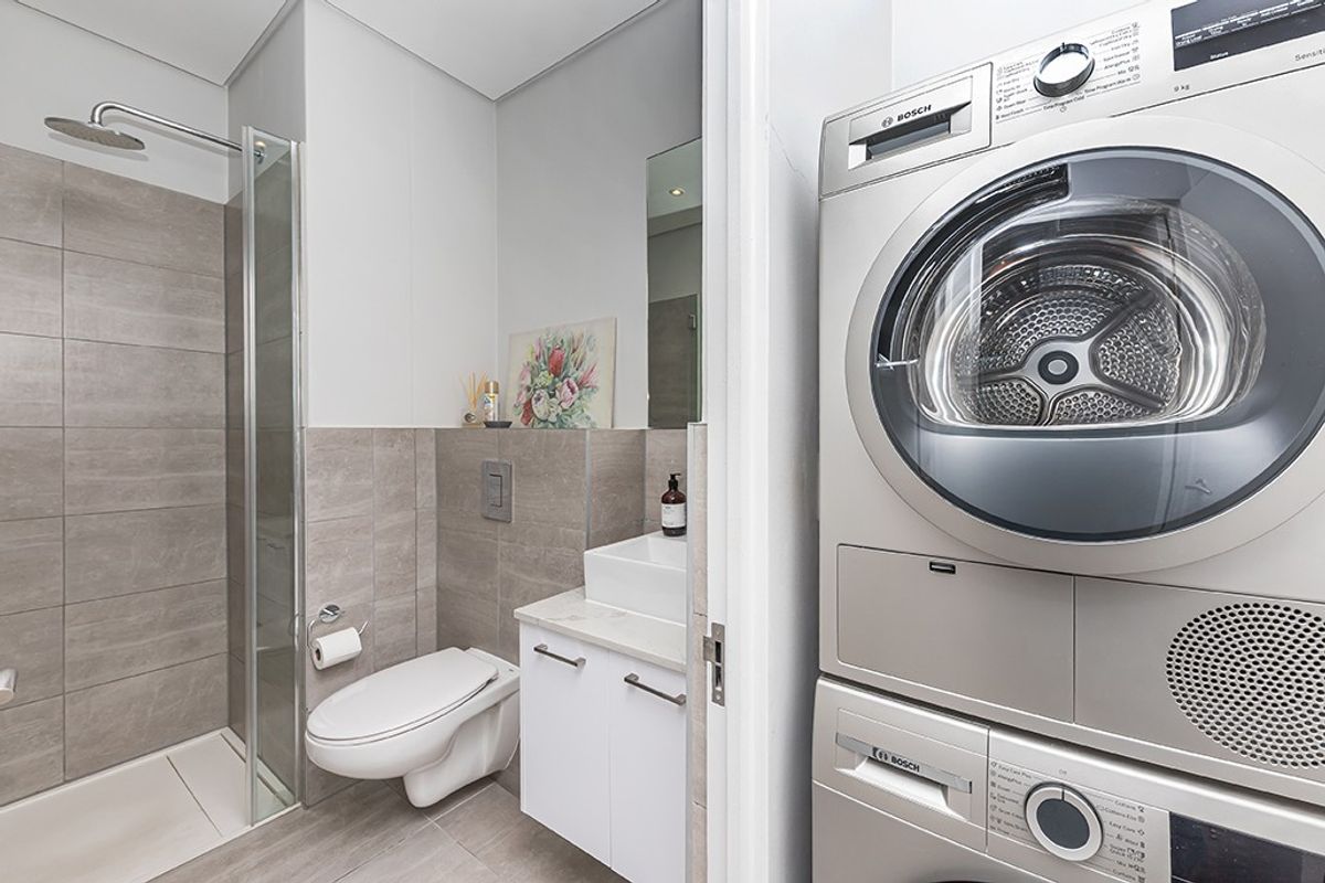 Modern bathroom featuring a walk-in shower, wall-hung toilet, and vanity, with an adjacent laundry area containing stacked washing machine and tumble dryer.