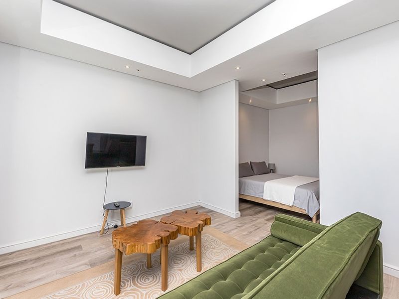 An open-plan living area featuring modern recessed ceiling details, integrated lighting, and light-toned laminate flooring, seamlessly connecting to a bedroom alcove.