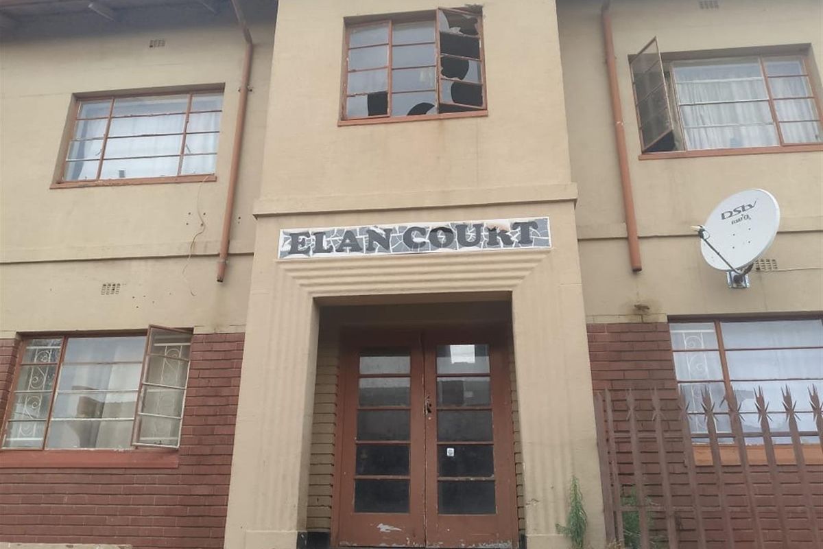 Front exterior view of Elan Court apartment building, featuring a central entrance and multi-pane windows showing signs of significant wear and damage.