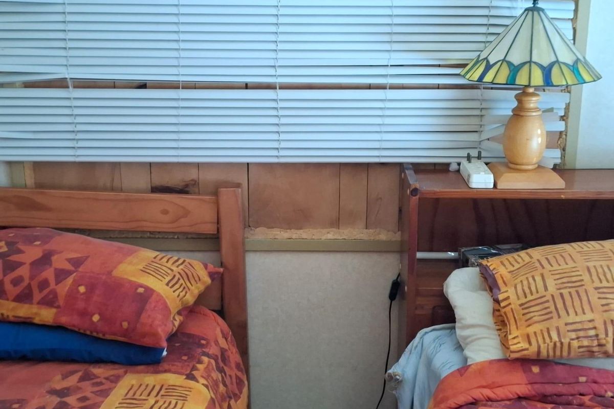 A bedroom featuring venetian blinds and visible damage to the wall paneling behind the bed area.