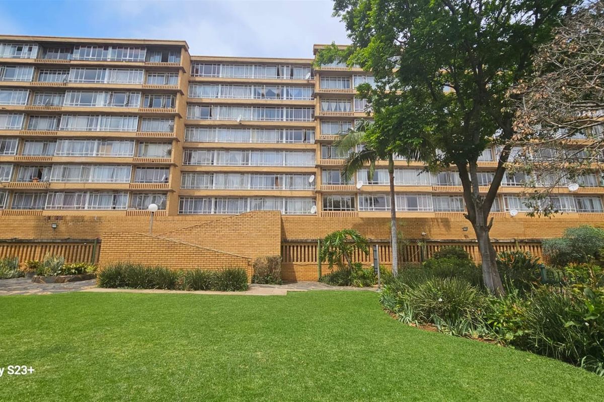A view of the rear facade of a multi-story apartment building featuring brick construction and extensive window glazing, set against a landscaped garden area.