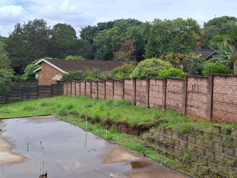 A vacant residential plot featuring a concrete slab foundation, brick boundary wall, and tiered concrete retaining wall with visible soil erosion.