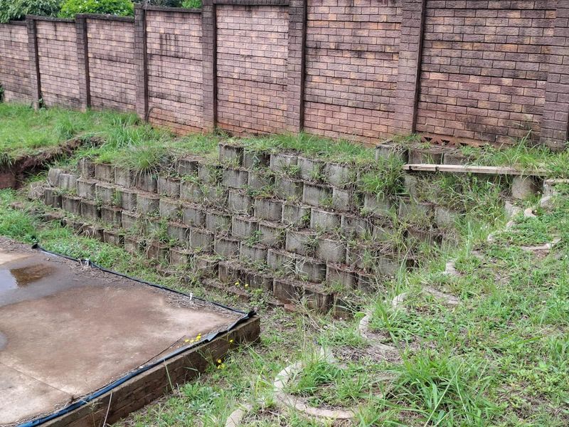 A residential garden area featuring a brick boundary wall, a tiered concrete block retaining wall, and a concrete slab foundation.