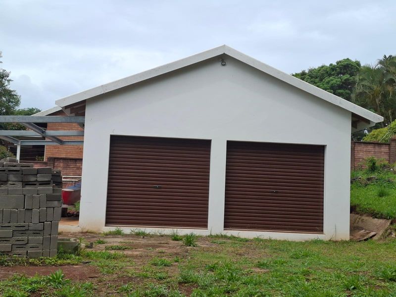 A detached double garage featuring brown roller doors and a pitched roof, situated on a residential plot with adjacent metal carport structure.