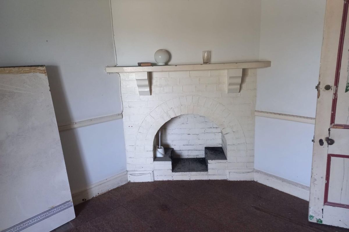 A corner room featuring a painted brick fireplace with a mantelpiece, worn carpeting, and a traditional timber door.