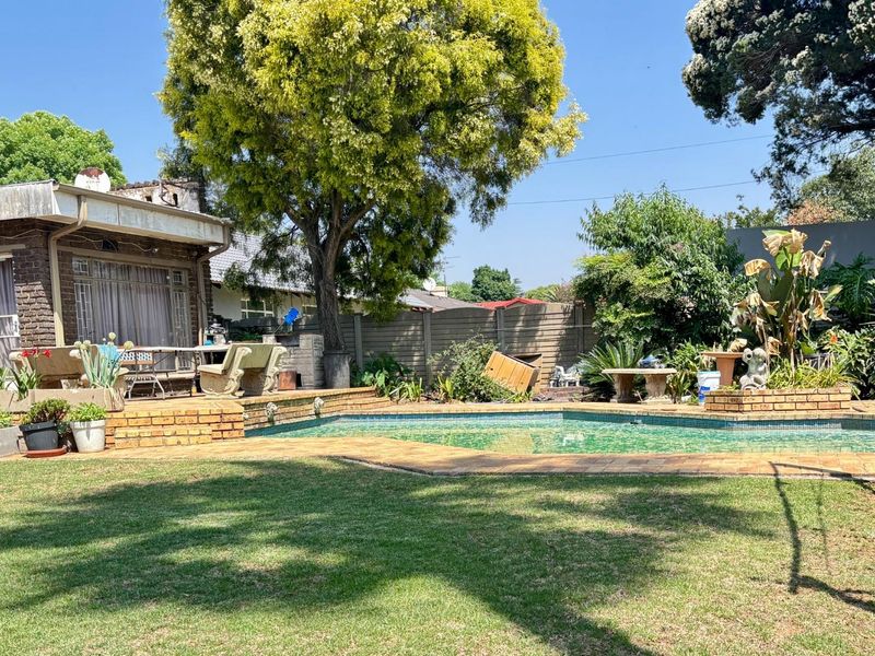 A backyard pool area featuring a swimming pool with brick surround, established garden, and a brick-walled house exterior.