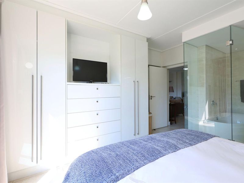 Modern bedroom featuring extensive built-in white cabinetry and a contemporary glass-enclosed en-suite bathroom with a soaking tub.