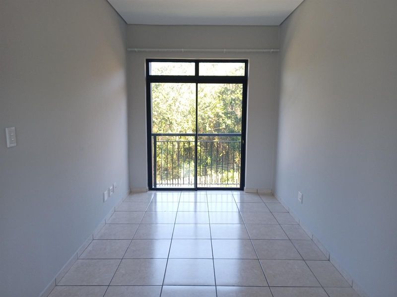 A bright, empty bedroom featuring neutral tiled flooring and a sliding glass door that opens onto a balcony with a leafy outlook.
