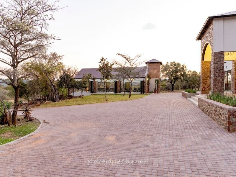 A view of the paved driveway and exterior grounds, featuring stone-clad architecture and a secondary building structure set within a landscaped small holding.