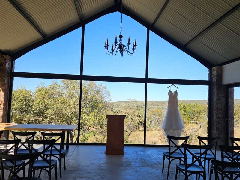 A bright, open-plan event space featuring high vaulted ceilings, exposed steel trusses, and expansive floor-to-ceiling glass walls offering panoramic views of the surrounding landscape.
