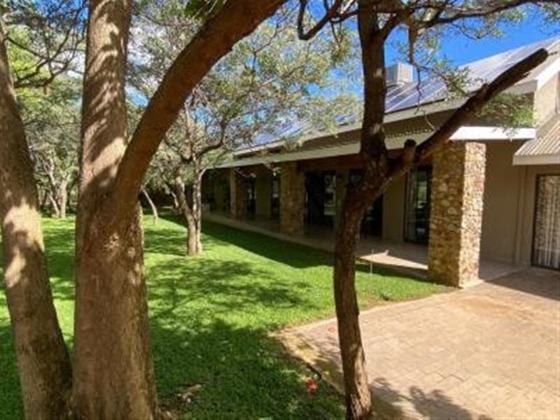 A spacious covered patio area featuring stone-clad pillars, overlooking a lush, tree-lined garden with paved pathways.