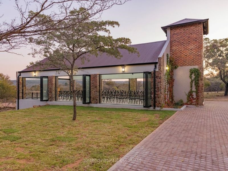 A modern event venue featuring exposed brickwork, large glass folding doors, and a metal roof, set within a landscaped small holding environment.