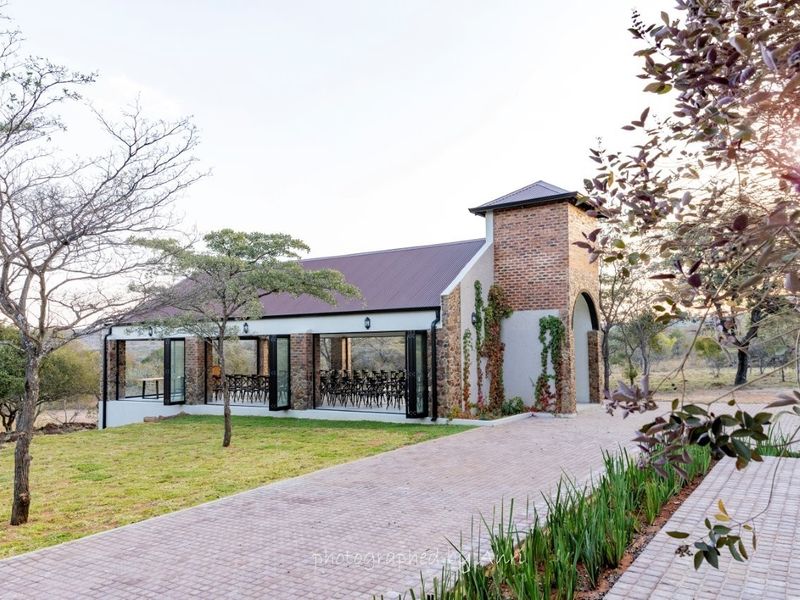 A charming brick-clad event venue or clubhouse featuring large glass folding doors, a metal roof, and a paved driveway set within a natural landscape.