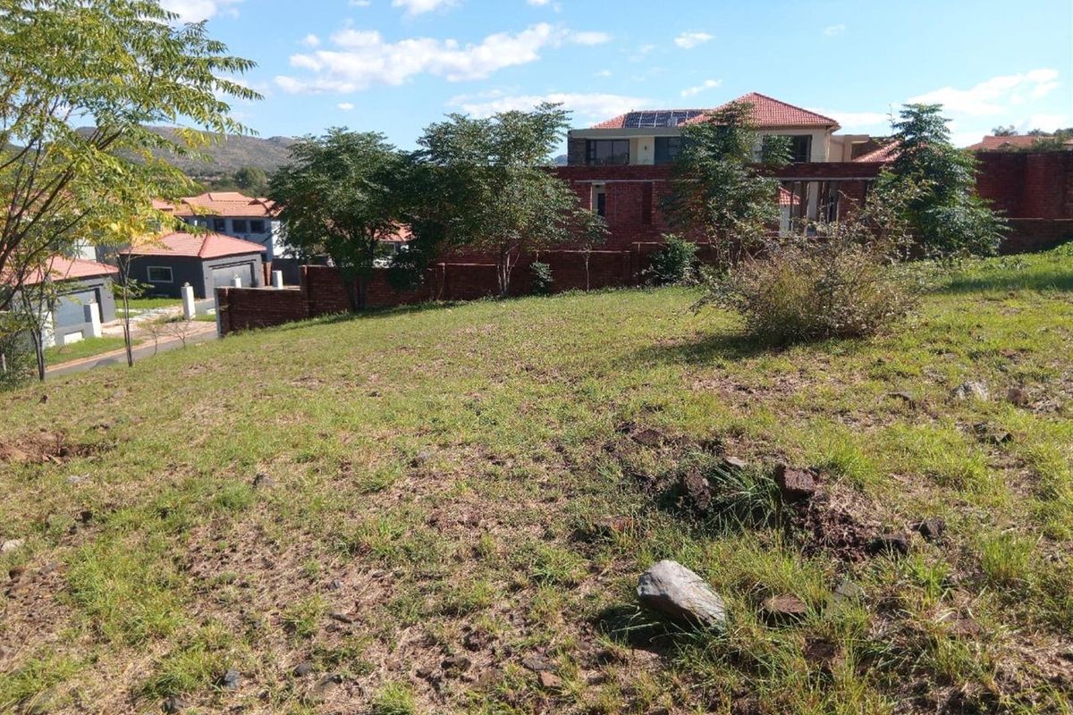 A view of a sloped vacant residential plot featuring established trees and a brick boundary wall in a residential estate setting.