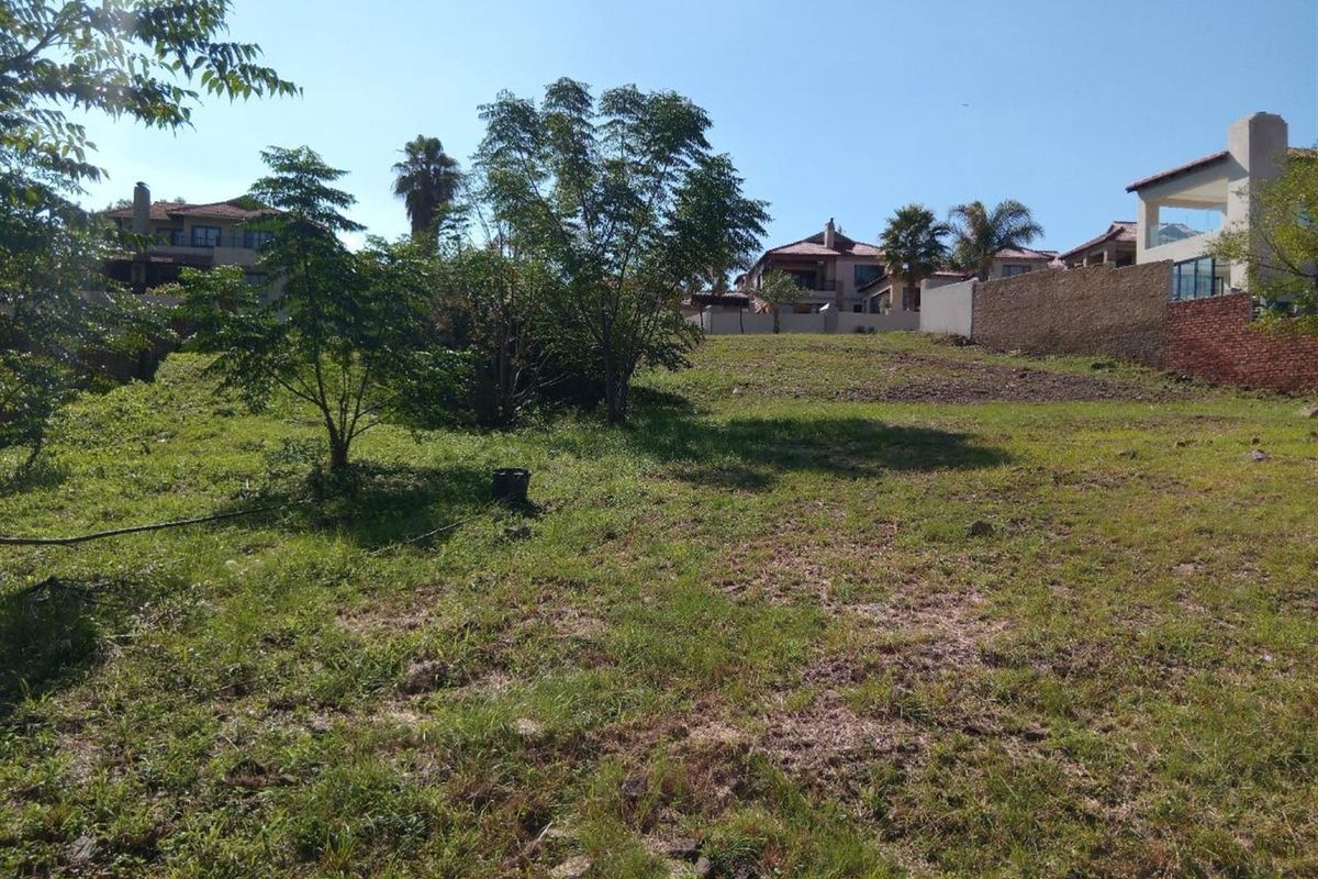A view of a vacant residential plot featuring established trees and a boundary wall, situated within a developed neighborhood.