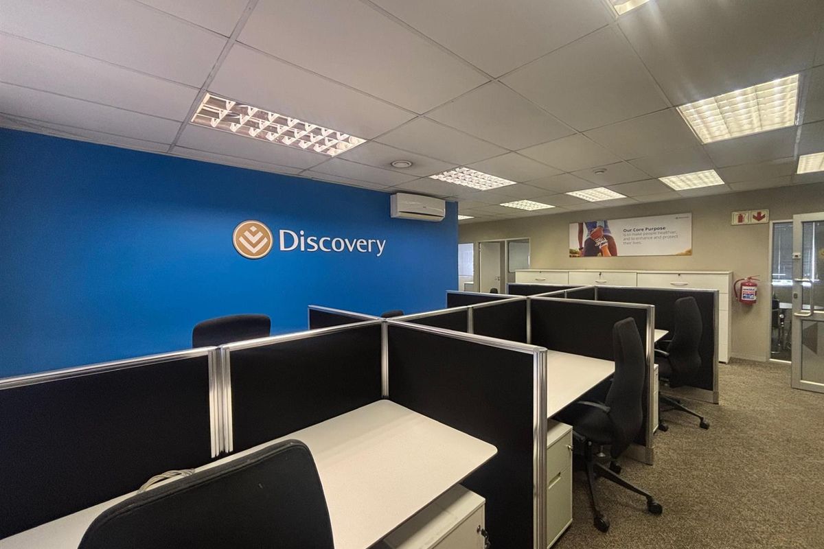 A professional office space featuring a suspended ceiling, recessed lighting, carpeted flooring, and integrated cubicle workstations with a branded feature wall.