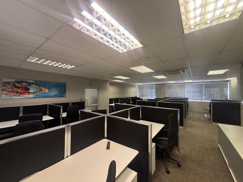 Open-plan office space featuring modular cubicle workstations, suspended ceilings with recessed lighting, and carpeted flooring with large windows and venetian blinds.