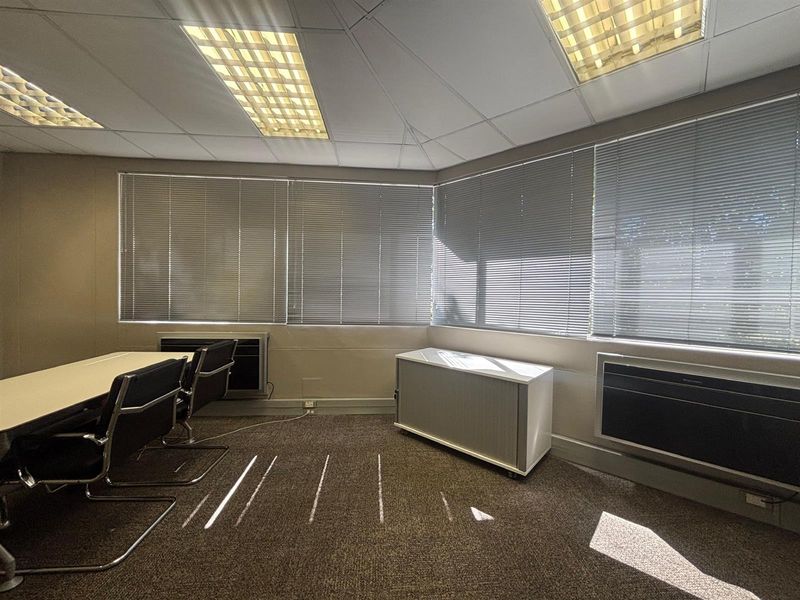 A functional office space featuring carpeted flooring, suspended ceilings with recessed lighting, wall-mounted air conditioning units, and large windows with fitted venetian blinds.