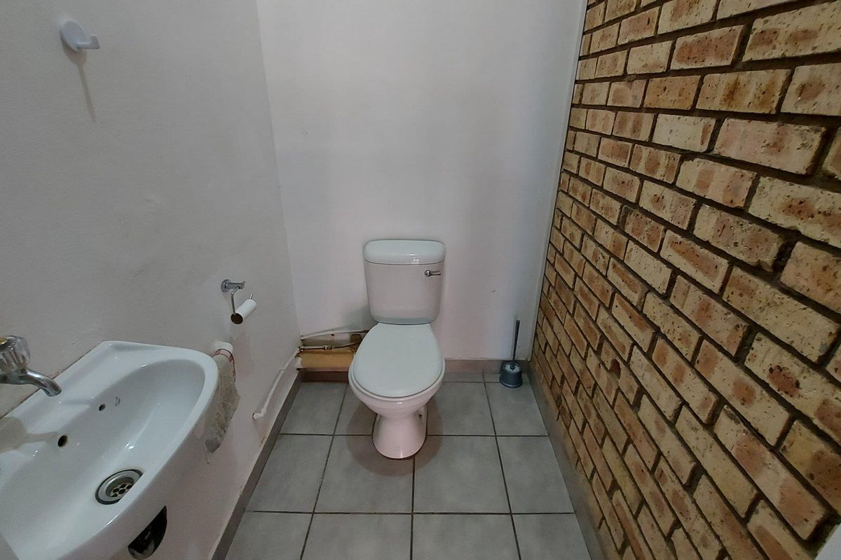 A compact commercial restroom featuring a white ceramic toilet, a wall-mounted basin, tiled flooring, and a decorative exposed brick feature wall.