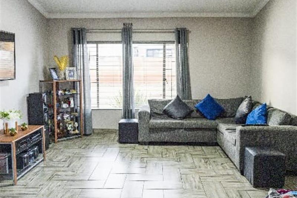 A spacious living room featuring modern herringbone-patterned floor tiles, neutral walls, and a large window with security bars.