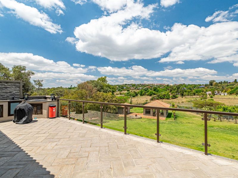 Spacious elevated patio featuring stone-look tiling, glass balustrades, and a built-in braai area, offering expansive views over the garden and surrounding landscape.