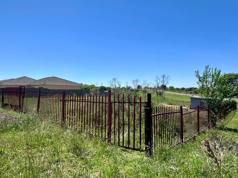 A vacant residential plot enclosed by metal palisade fencing, featuring overgrown vegetation and clear views of the surrounding neighborhood.