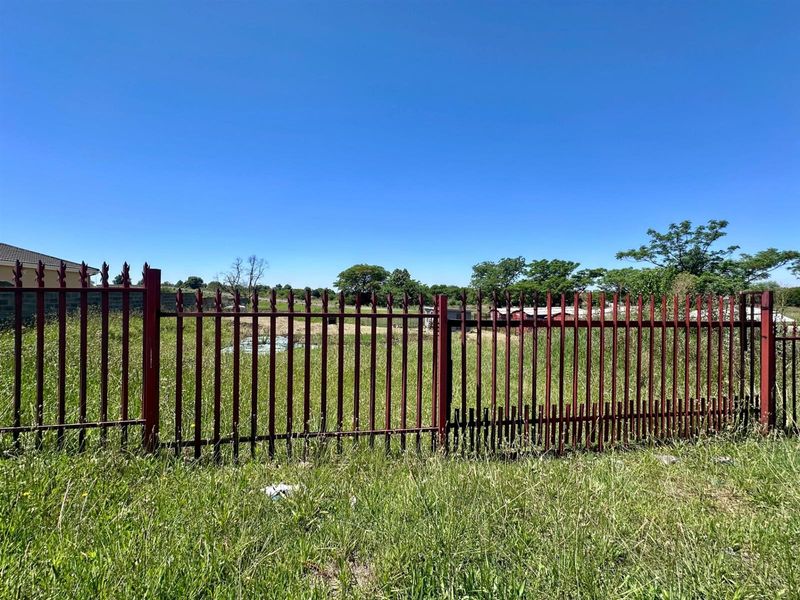 A view of a vacant residential plot enclosed by metal palisade fencing, showing overgrown grass and a damaged section of the perimeter fence.