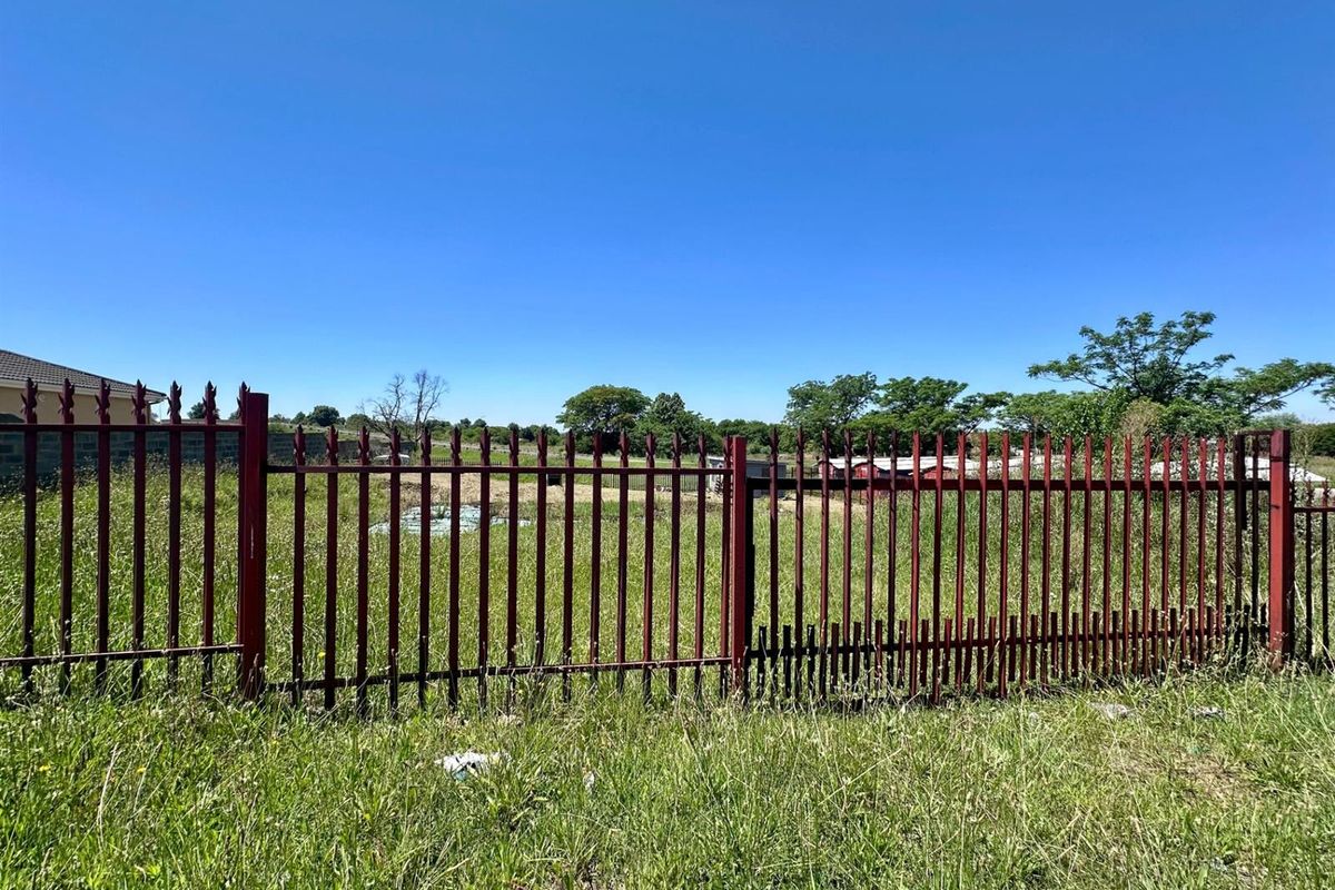 A view of a vacant residential plot enclosed by metal palisade fencing, showing overgrown grass and a damaged section of the perimeter fence.