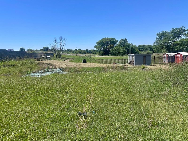 A view of a vacant residential plot featuring overgrown grass, perimeter metal fencing, and a small storage shed on the property.