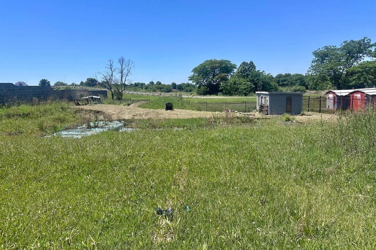 A view of a vacant residential plot featuring overgrown grass, perimeter metal fencing, and a small storage shed on the property.