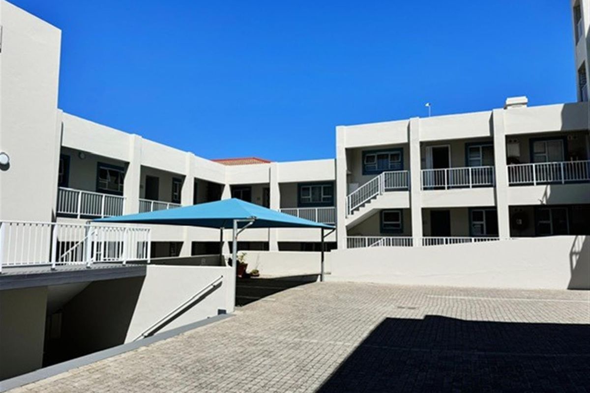 Exterior view of the apartment complex featuring paved courtyard, multi-level walkways with metal railings, and a shaded parking area.