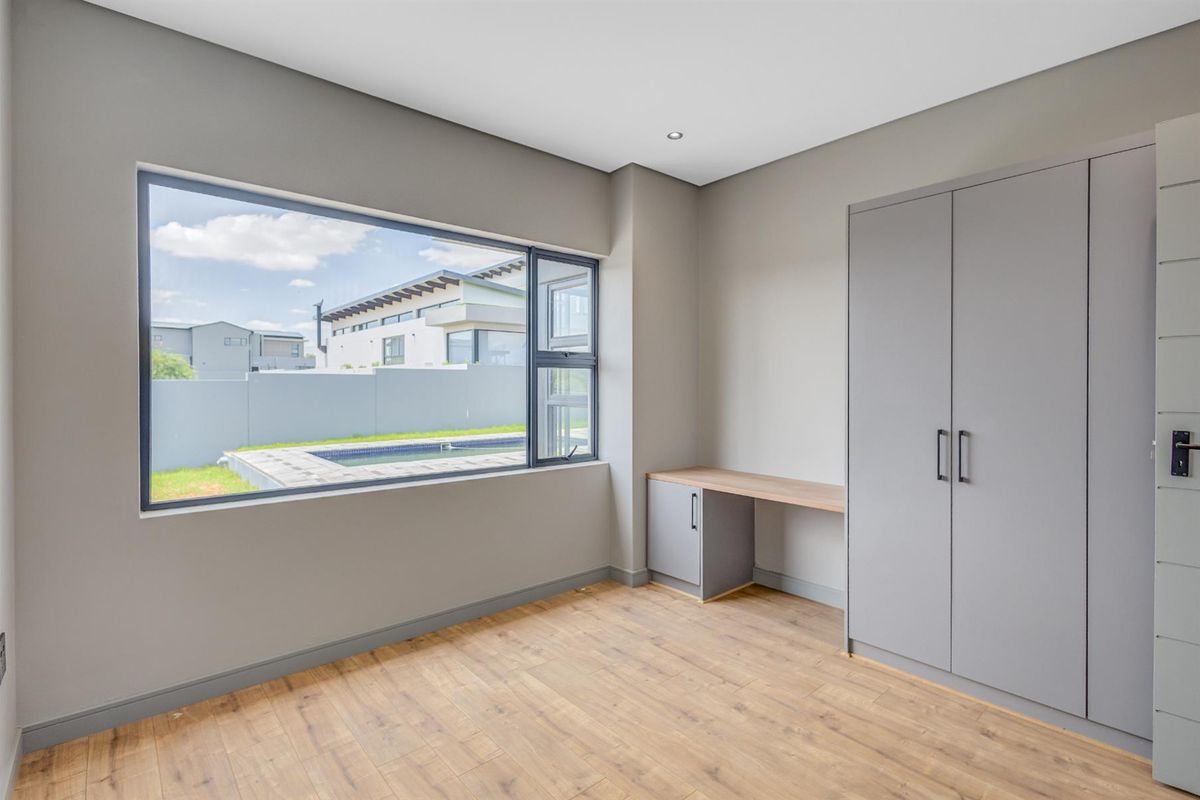 A bright, modern bedroom featuring wood-look flooring, built-in grey cabinetry with an integrated desk, and a large window overlooking the pool area.