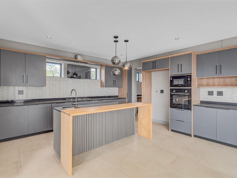 Modern kitchen featuring grey cabinetry, light wood accents, a central island with breakfast bar, built-in oven, microwave, and tiled backsplash.