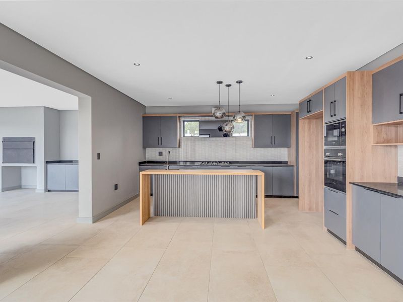 Modern open-plan kitchen featuring sleek grey cabinetry, wood-tone accents, integrated oven, and a central island, seamlessly flowing into an adjacent utility area.