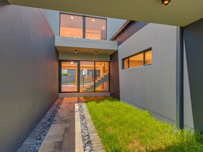 A modern architectural entrance featuring a paved walkway, landscaped garden strip, and large glass facade with a visible staircase.