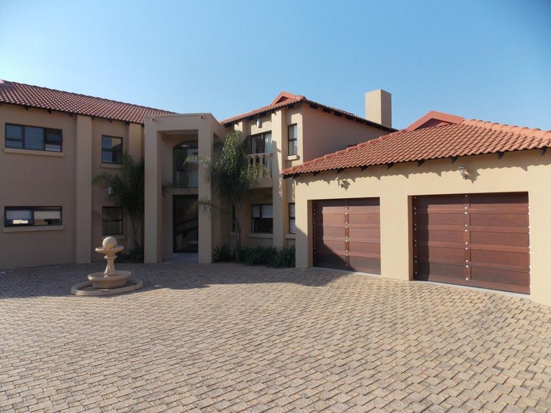 A well-maintained double-story house featuring a paved driveway, double garage with wooden doors, and a decorative stone fountain in the front courtyard.