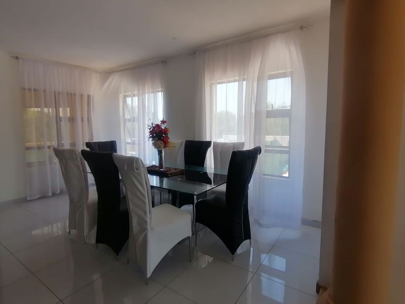 A bright dining room featuring large windows with sheer curtains and polished floor tiles, creating a clean and airy atmosphere.