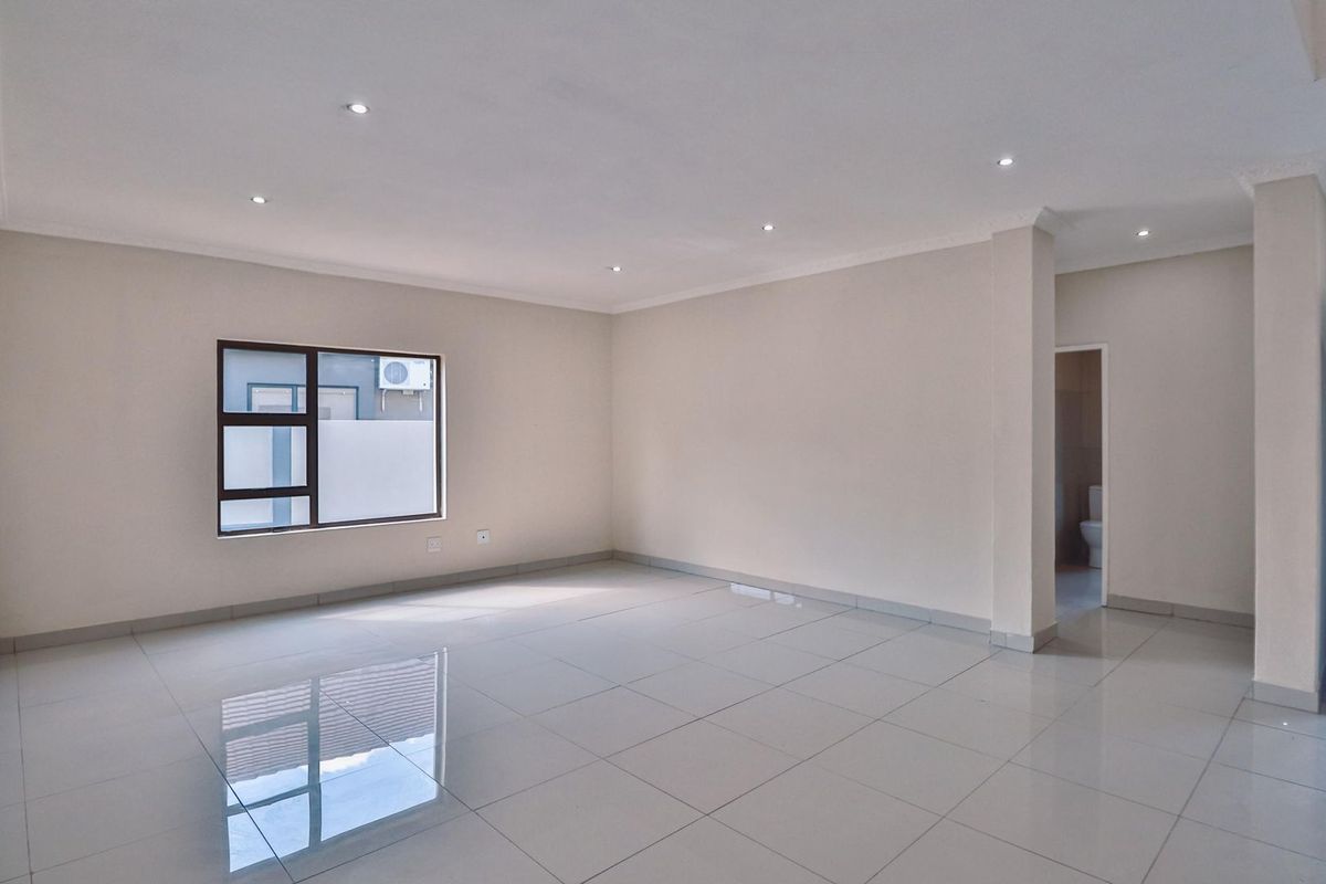 A spacious, empty living area featuring neutral tiled flooring, recessed downlighting, and a large window, with an adjacent doorway leading to a bathroom.