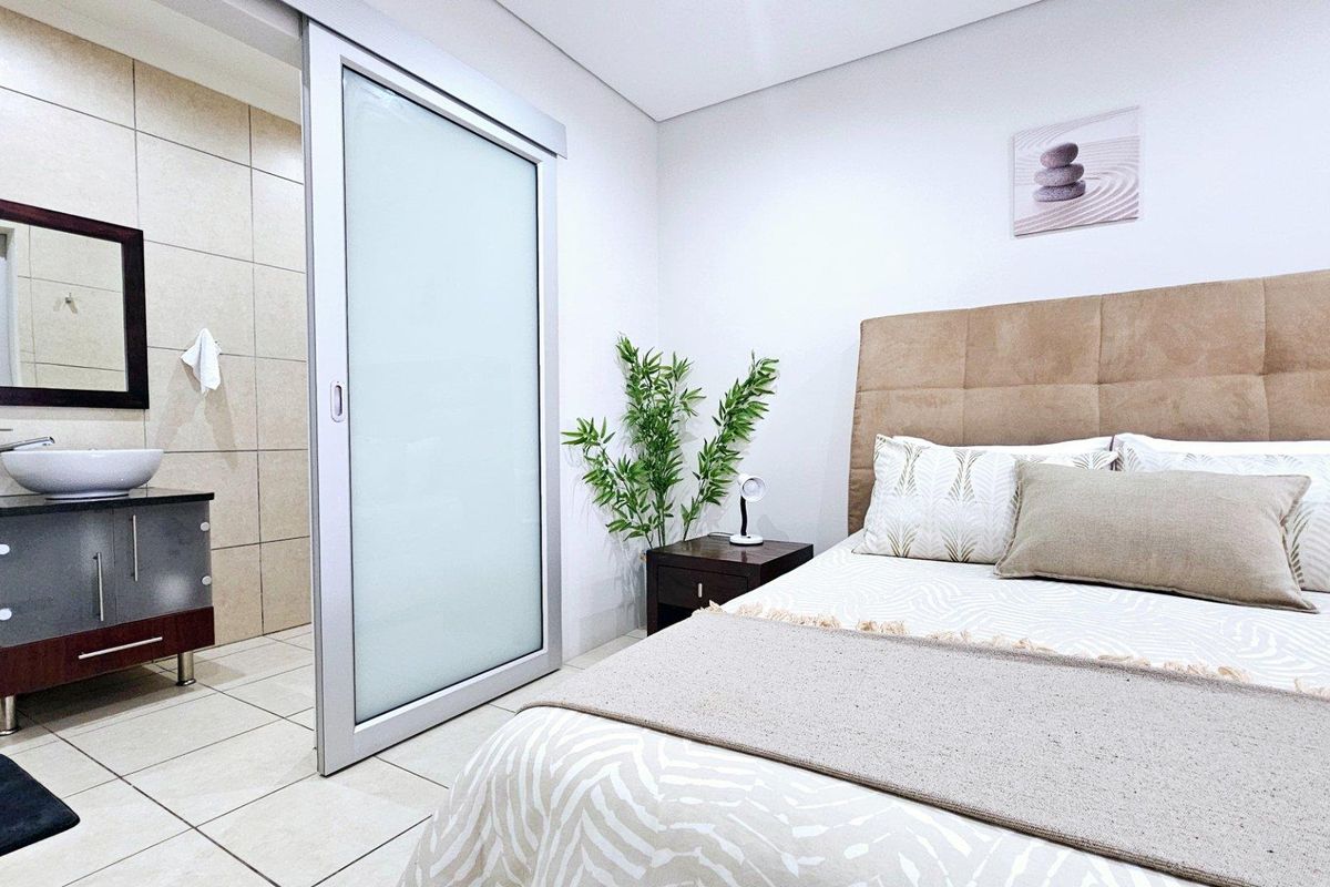 A modern bedroom featuring an en-suite bathroom area with a vessel sink, vanity cabinet, and a sliding glass door partition.