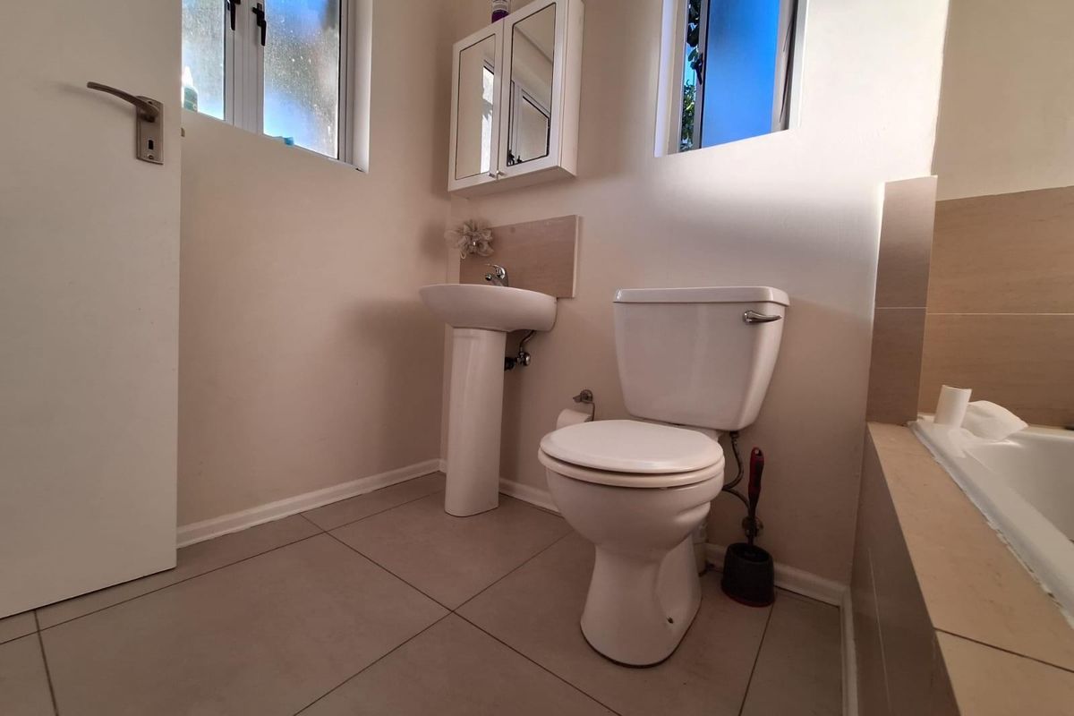 A functional bathroom featuring a pedestal basin, toilet, and tiled bath surround with neutral wall finishes and natural light from two windows.