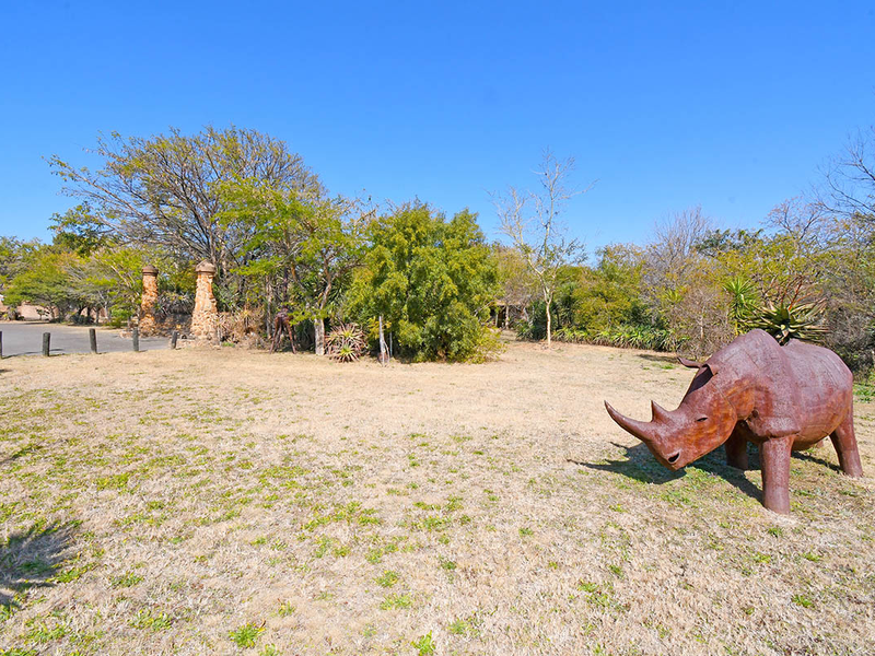 A spacious, well-maintained garden area featuring mature trees, stone pillar accents, and a clear driveway approach within a natural lodge setting.