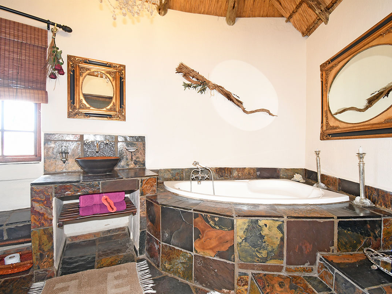 A rustic bathroom featuring a built-in corner bathtub and stone-clad vanity with a vessel sink, set under a traditional thatched roof.
