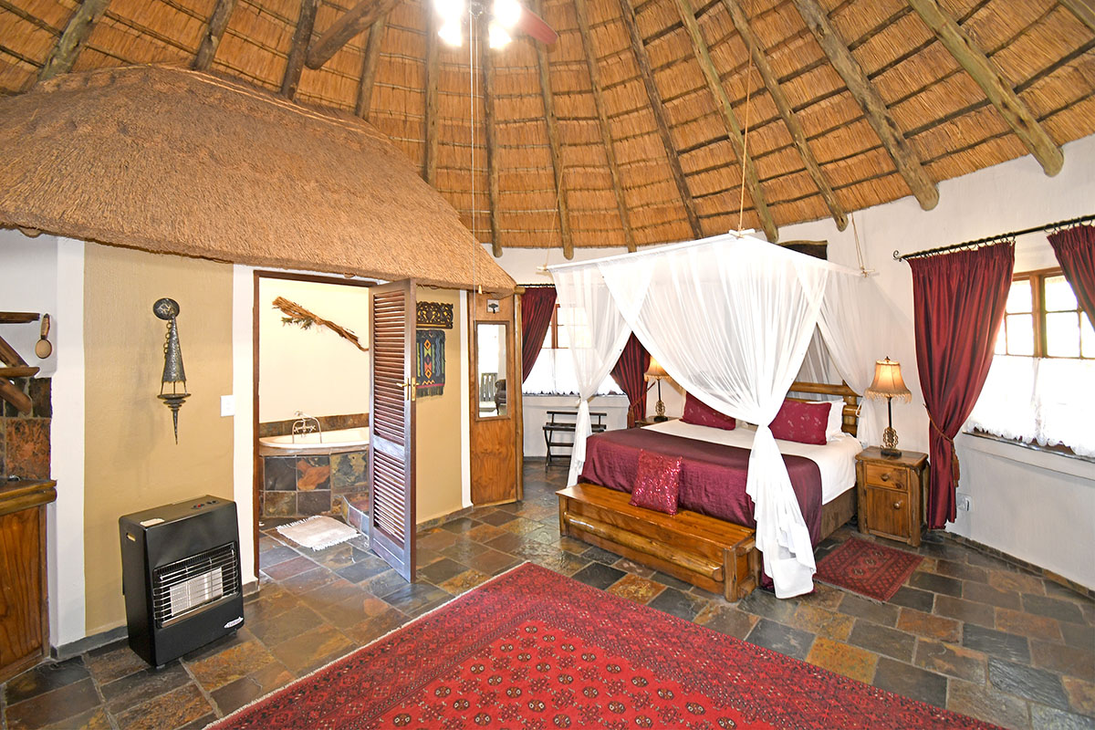 Spacious bedroom in a lodge-style unit featuring a traditional thatched roof, exposed timber rafters, slate flooring, and an integrated en-suite bathroom.