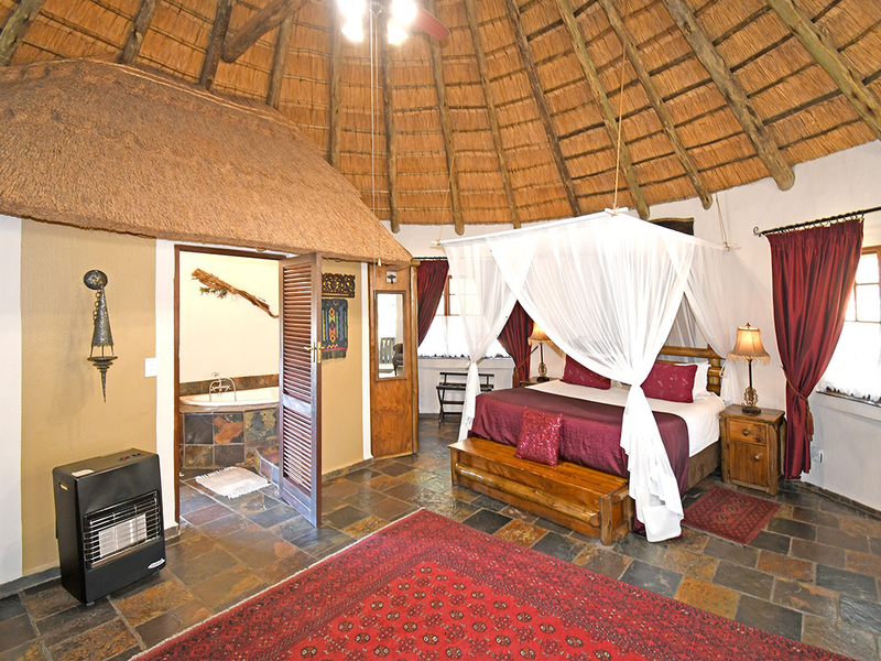 Spacious bedroom in a lodge-style unit featuring a traditional thatched roof, exposed timber rafters, slate flooring, and an integrated en-suite bathroom.