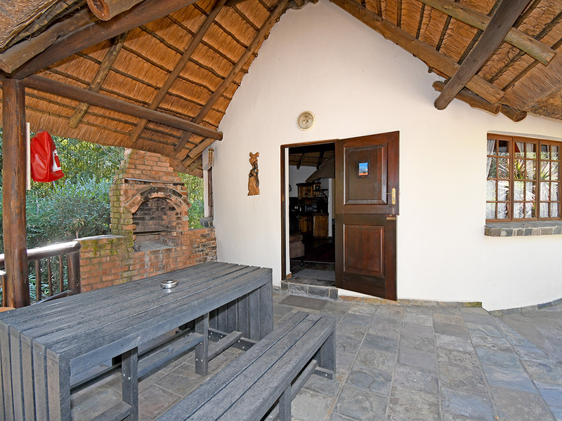 Covered outdoor patio featuring a traditional thatched roof, exposed timber rafters, built-in brick braai, and durable slate tile flooring.