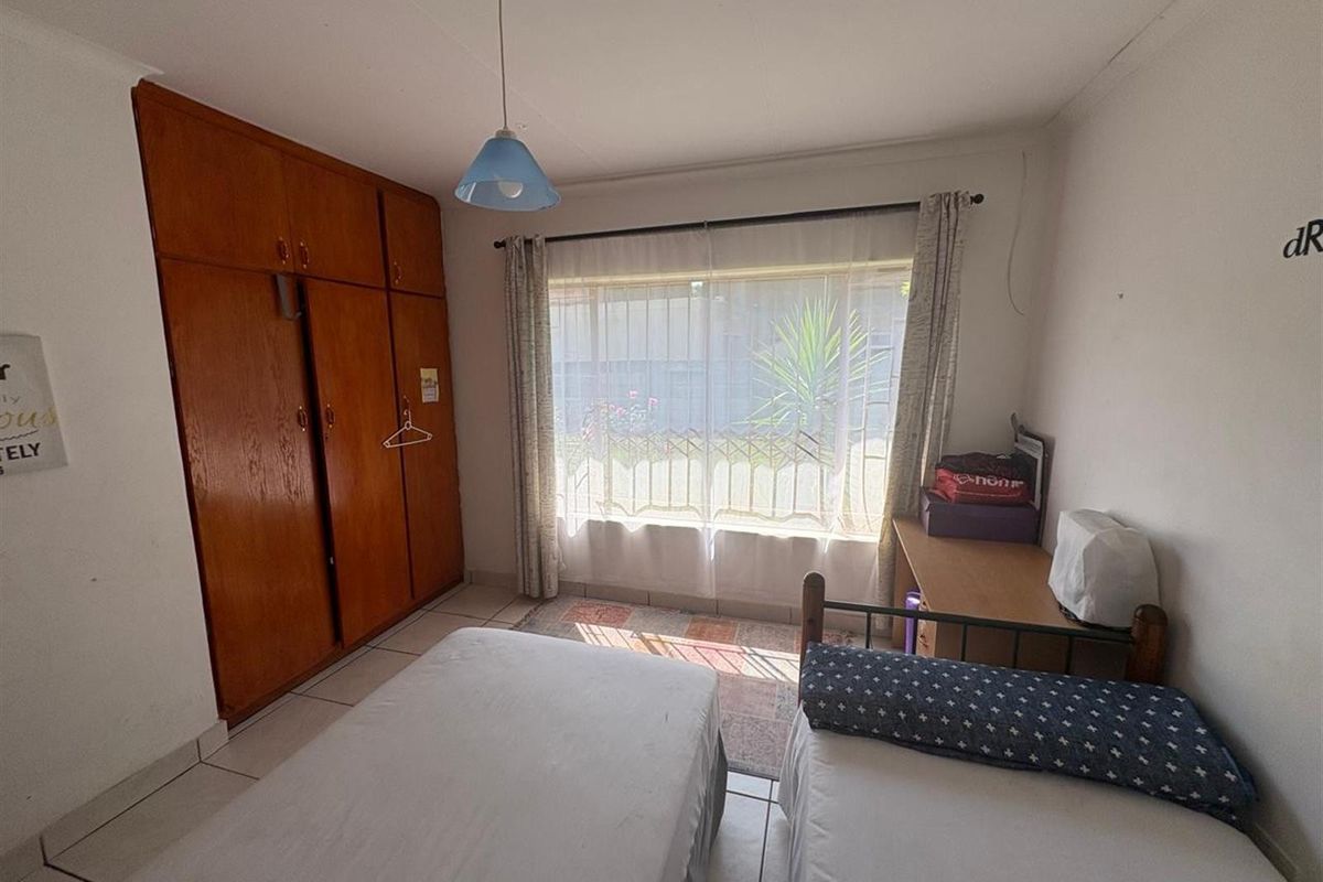 A functional bedroom featuring built-in wooden cupboards, tiled flooring, and a large window allowing for natural light.