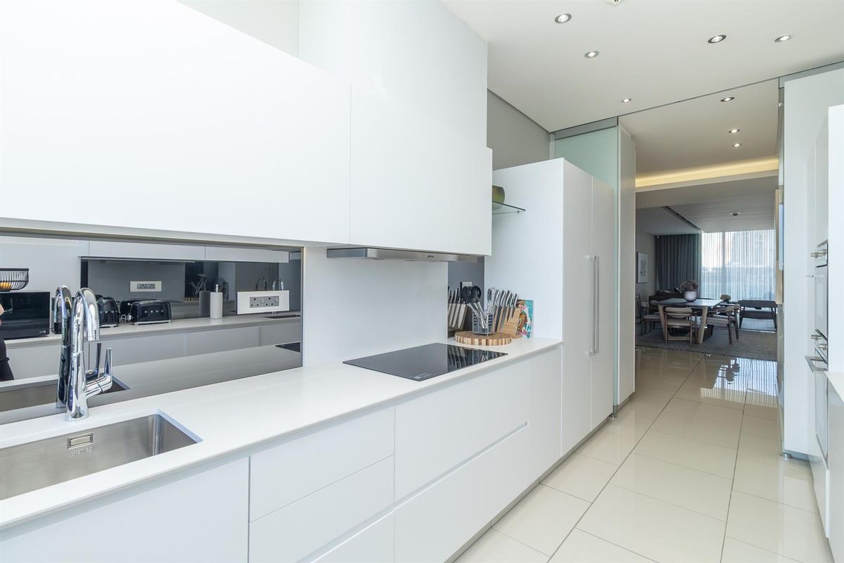 Modern, sleek kitchen featuring white handleless cabinetry, integrated appliances, a glass splashback, and polished floor tiles, seamlessly flowing into the adjacent living area.