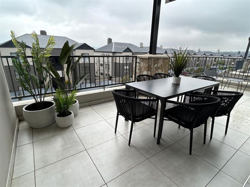 A spacious covered balcony featuring modern tiled flooring, a stone-clad support pillar, and a secure metal balustrade overlooking the surrounding residential complex.
