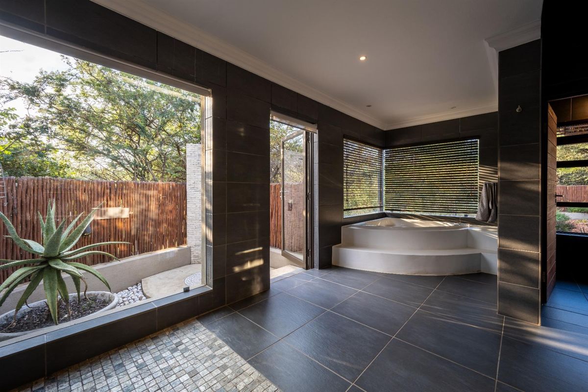 Modern bathroom featuring a built-in corner bathtub, dark large-format tiling, and expansive windows providing natural light and garden views.